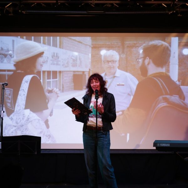 performer on stage with book in hand, screen behingd showing a picture of three figures as Darlington station