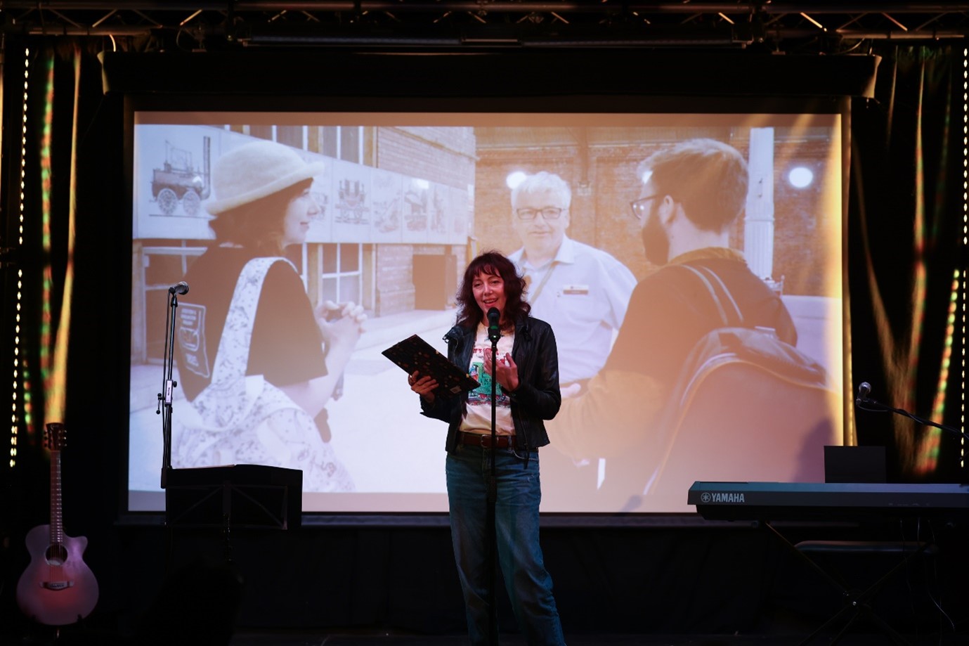 performer on stage with book in hand, screen behingd showing a picture of three figures as Darlington station