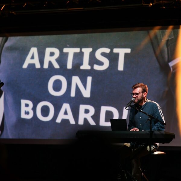 Performer on stage wearing blue shirt, playing keyboard, on screen behind is a picture saying artist on board