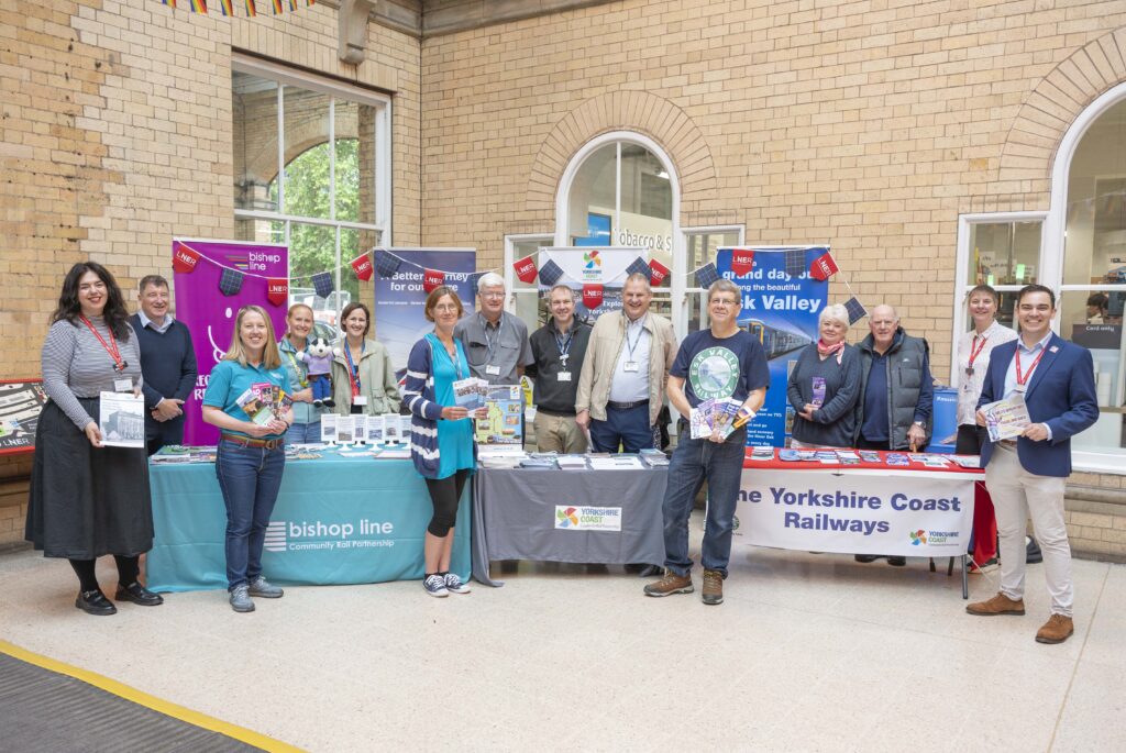 Group of 14 people standing inside a station building with rectangular and arched windows in the background. The people are holding leaflets. 5 of the people are standing in front of 3 tables each with a table cloth on. One table cloth shows the Bishop Line CRP logo, the second table shows the Yorkshire Coast CRP logo and the third table cloth has a banner reading The Yorkshire Coast Railways and features the Yorkshire Coast CRP logo and the Esk Valley Railway logo. On the tables there are many leaflets and promotional merchandise. 9 of the people are standing behind the tables, one person is holding a soft toy badger. There are 4 pull up banners behind them with bunting hanging from it. The bunting features the LNER logo and the LNER tartan.