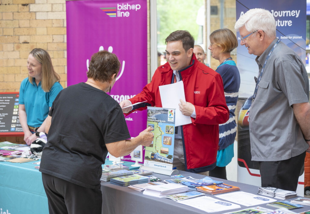 Photo of 6 adults, one in front of a table with leaflets on, three people behind the table talking to people in front of the table, and two further people talking in the back ground. In the back of the image are two pull up banners one featuring the Bishop Line logo and the other featuring an LNER train.