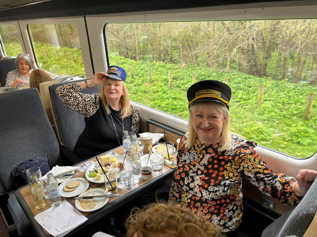 Photo of 2 ladies sitting at a table inside a Weardale Railway special cheese and gin train, the table is covered with plates and glasses. The ladies are wearing railway hats.